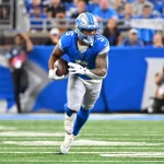 FootballR - NFL - Ein Footballspieler in blauem Trikot und Helm, ähnlich wie David Montgomery, läuft mit dem Ball über ein Feld. Er ist konzentriert und marschiert, im unscharfen Hintergrund sind eine Menge und ein Fotograf zu sehen. Das Feld hat weiße Yard-Linien und Kunstrasen. Diese Beschreibung wurde automatisch generiert.