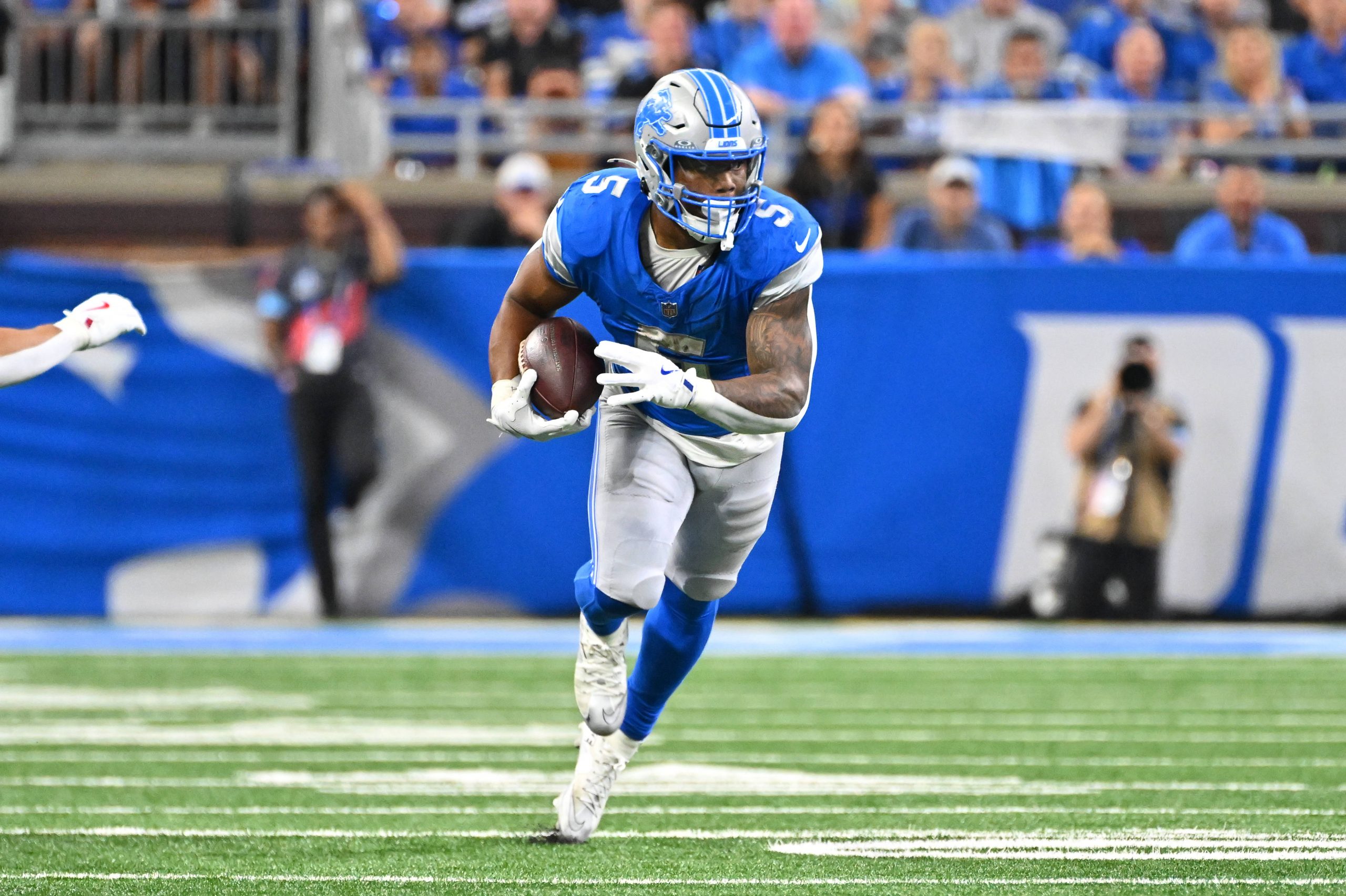FootballR - NFL - Ein Footballspieler in blauem Trikot und Helm, ähnlich wie David Montgomery, läuft mit dem Ball über ein Feld. Er ist konzentriert und marschiert, im unscharfen Hintergrund sind eine Menge und ein Fotograf zu sehen. Das Feld hat weiße Yard-Linien und Kunstrasen. Diese Beschreibung wurde automatisch generiert.