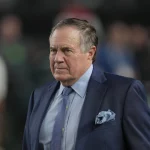 FootballR - NFL - Vince Lombardi Trophy - Ein Mann in blauem Anzug und Krawatte steht im Freien und strahlt Kritik aus. Mit seinen kurzen grauen Haaren und dem konzentrierten Gesichtsausdruck erinnert er an Bill Belichick. Ein Einstecktuch ziert sein Jackett. Der unscharfe Hintergrund deutet auf eine Veranstaltung hin, möglicherweise mit Beteiligung von Jets-Besitzer oder ähnlichen Figuren. Diese Beschreibung wurde automatisch generiert.