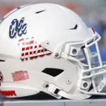 FootballR - NFL - Ein weißer Footballhelm mit dem ikonischen Ole Miss Logo in roter und blauer Schreibschrift an der Seite ist zu sehen. Er hat eine glatte weiße Gesichtsmaske, einen Aufkleber mit der amerikanischen Flagge und ein SEC-Logo auf der Rückseite, alles vor einem verschwommenen Hintergrund. Diese Beschreibung wurde automatisch generiert.