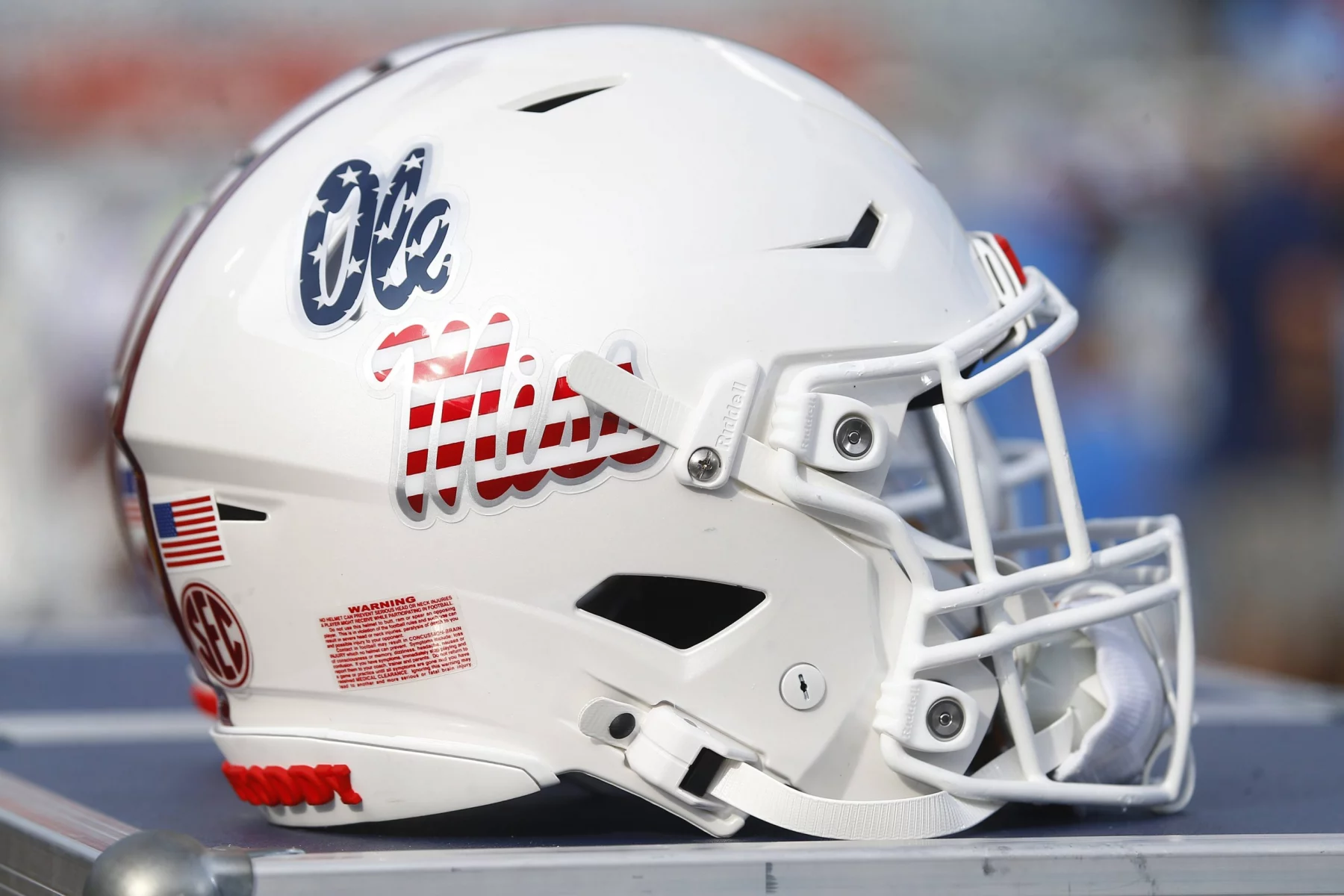 FootballR - NFL - Ein weißer Footballhelm mit dem ikonischen Ole Miss Logo in roter und blauer Schreibschrift an der Seite ist zu sehen. Er hat eine glatte weiße Gesichtsmaske, einen Aufkleber mit der amerikanischen Flagge und ein SEC-Logo auf der Rückseite, alles vor einem verschwommenen Hintergrund. Diese Beschreibung wurde automatisch generiert.