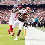 FootballR - NFL - Amari Cooper in braun-orangefarbener Uniform fängt einen Pass in der Nähe der Seitenlinie, dicht gefolgt von einem Verteidiger der Bills in weiß-blauer Uniform. Beide Spieler stehen auf einem grünen Feld, während eine große Menschenmenge auf den Tribünen des Stadions das Geschehen verfolgt. Diese Beschreibung wurde automatisch generiert.
