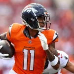 FootballR - NFL - Broncos-Receiver Josh Reynolds im orangefarbenen Denver Broncos-Trikot mit der Nummer 11 rennt über das Feld und hält dabei einen Football in seiner rechten Hand. Während er schnell einem Spieler im weißen Trikot ausweicht, wandert sein Blick nach links. Der unscharfe Hintergrund fängt die rasante Action des Augenblicks ein. Diese Beschreibung wurde automatisch generiert.