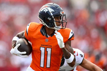 FootballR - NFL - Broncos-Receiver Josh Reynolds im orangefarbenen Denver Broncos-Trikot mit der Nummer 11 rennt über das Feld und hält dabei einen Football in seiner rechten Hand. Während er schnell einem Spieler im weißen Trikot ausweicht, wandert sein Blick nach links. Der unscharfe Hintergrund fängt die rasante Action des Augenblicks ein. Diese Beschreibung wurde automatisch generiert.