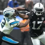 FootballR - NFL - Davante Adams, in schwarz-weißer Uniform und Helm, streckt einen gegnerischen Spieler in weiß-blaugrün-schwarzer Uniform und Helm mit ausgestrecktem Arm aus, der ihn während des Spiels zu tackeln versucht. Adams hält den Football fest, während andere Figuren und ein verschwommener Hintergrund auf Stadionaktivität hinweisen. Diese Beschreibung wurde automatisch generiert.