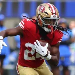 FootballR - NFL Power Ranking - Ein Footballspieler in rot-goldener Uniform mit der Nummer 24 stürmt während eines Spiels entschlossen nach vorne. Auf seinem goldenen Helm ist das Logo der San Francisco 49ers zu sehen, das ihn als Jordan Mason kennzeichnet. Teamkollegen und verschwommene Zuschauer der Seahawks bilden die dynamische Kulisse. Diese Beschreibung wurde automatisch generiert.
