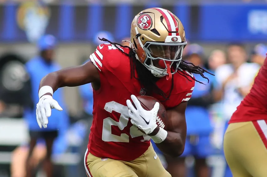 FootballR - NFL Power Ranking - Ein Footballspieler in rot-goldener Uniform mit der Nummer 24 stürmt während eines Spiels entschlossen nach vorne. Auf seinem goldenen Helm ist das Logo der San Francisco 49ers zu sehen, das ihn als Jordan Mason kennzeichnet. Teamkollegen und verschwommene Zuschauer der Seahawks bilden die dynamische Kulisse. Diese Beschreibung wurde automatisch generiert.