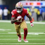 FootballR - NFL - Brandon Aiyuk sprintet in einer rot-goldenen Uniform über das grüne Feld. Er trägt einen Helm mit Gesichtsschutz, rote Handschuhe und weiße Stollenschuhe. Verschwommene Zuschauer verfolgen das Geschehen, während die Anzeigetafel in der oberen rechten Ecke ein laufendes Spiel anzeigt. Diese Beschreibung wurde automatisch generiert.