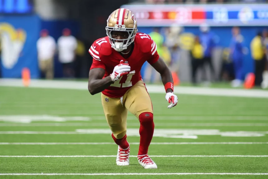 FootballR - NFL - Brandon Aiyuk sprintet in einer rot-goldenen Uniform über das grüne Feld. Er trägt einen Helm mit Gesichtsschutz, rote Handschuhe und weiße Stollenschuhe. Verschwommene Zuschauer verfolgen das Geschehen, während die Anzeigetafel in der oberen rechten Ecke ein laufendes Spiel anzeigt. Diese Beschreibung wurde automatisch generiert.