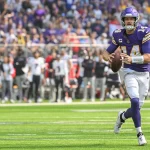 FootballR - NFL - Ein Footballspieler in lila-weißer Uniform mit der Nummer 14 hält einen Football und läuft über eine Wiese. Das klare Visier seines Helms glitzert in der Sonne und er imitiert die Beweglichkeit von Quarterback Sam Darnold. Im Hintergrund säumen Zuschauer und Teammitglieder in schwarz-weißer Kleidung die Seitenlinien, wie im Duell-Stadion. Diese Beschreibung wurde automatisch generiert.