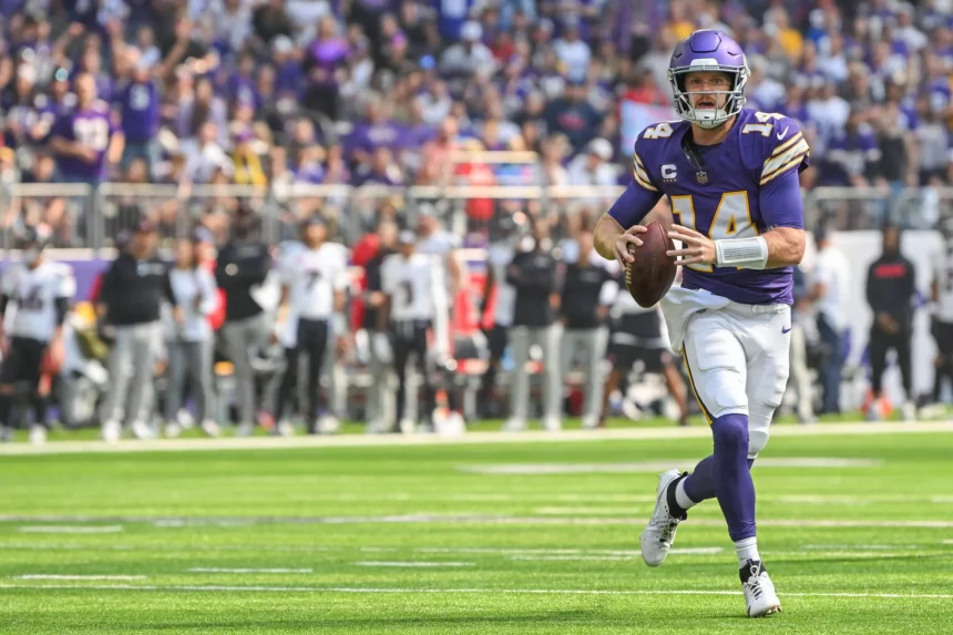 FootballR - NFL - Ein Footballspieler in lila-weißer Uniform mit der Nummer 14 hält einen Football und läuft über eine Wiese. Das klare Visier seines Helms glitzert in der Sonne und er imitiert die Beweglichkeit von Quarterback Sam Darnold. Im Hintergrund säumen Zuschauer und Teammitglieder in schwarz-weißer Kleidung die Seitenlinien, wie im Duell-Stadion. Diese Beschreibung wurde automatisch generiert.