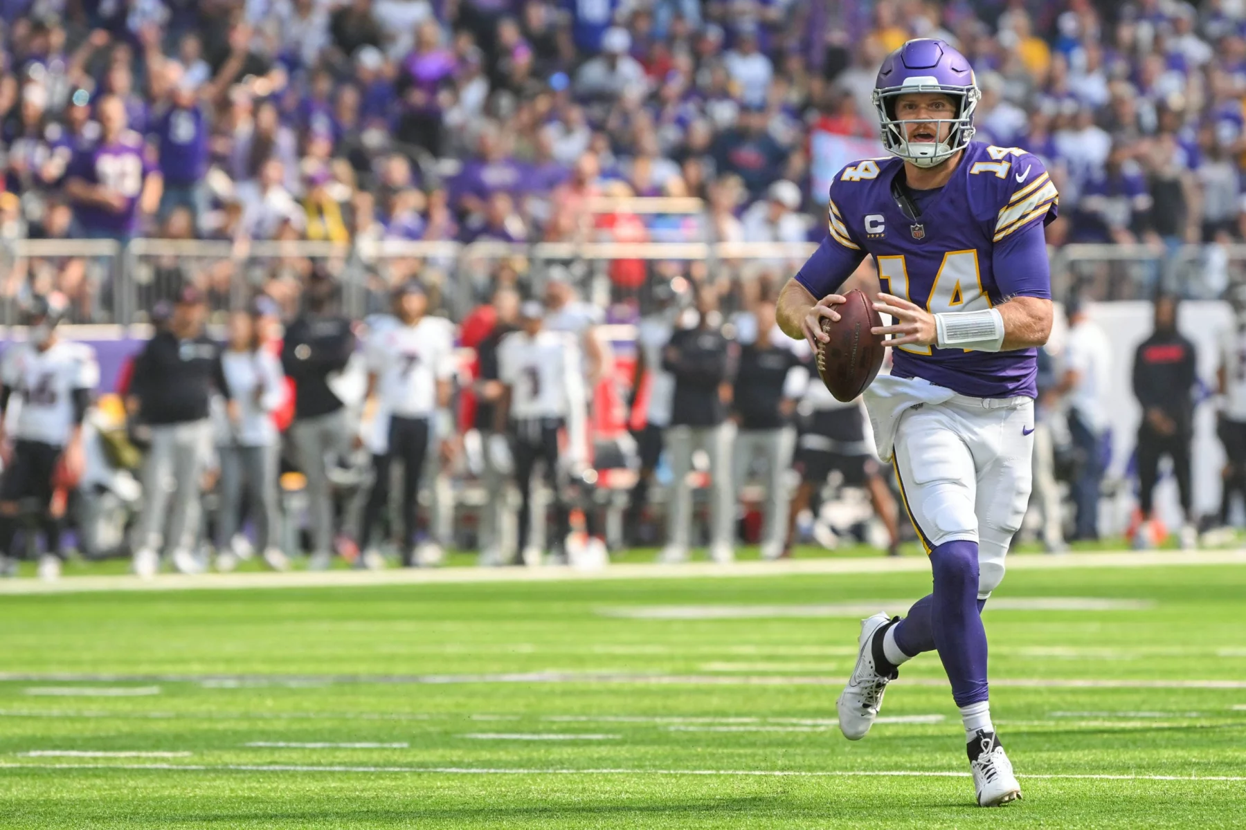 FootballR - NFL - Ein Footballspieler in lila-weißer Uniform mit der Nummer 14 hält einen Football und läuft über eine Wiese. Das klare Visier seines Helms glitzert in der Sonne und er imitiert die Beweglichkeit von Quarterback Sam Darnold. Im Hintergrund säumen Zuschauer und Teammitglieder in schwarz-weißer Kleidung die Seitenlinien, wie im Duell-Stadion. Diese Beschreibung wurde automatisch generiert.