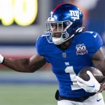 FootballR - NFL - Ein Footballspieler in einem blauen Giants-Trikot mit der Nummer 1 und einem speziellen 100-Saisons-Aufnäher sprintet über das Feld und hält den Ball fest umklammert. Mit seinem blauen Helm, Gesichtsschutz und weißen Handschuhen stürmt Malik Nabers nach vorne, während verschwommene Spieler und eine beleuchtete Anzeigetafel den Hintergrund füllen. Diese Beschreibung wurde automatisch generiert.