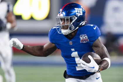 FootballR - NFL - Ein Footballspieler in einem blauen Giants-Trikot mit der Nummer 1 und einem speziellen 100-Saisons-Aufnäher sprintet über das Feld und hält den Ball fest umklammert. Mit seinem blauen Helm, Gesichtsschutz und weißen Handschuhen stürmt Malik Nabers nach vorne, während verschwommene Spieler und eine beleuchtete Anzeigetafel den Hintergrund füllen. Diese Beschreibung wurde automatisch generiert.