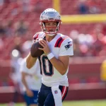 Wild Card - Woche 15 - NFL - Ein Footballspieler, Drake Maye, trägt ein weiß-rotes Trikot mit der Nummer 10 und einen Helm. Vor einer verschwommenen Stadionkulisse voller eifriger Zuschauer und hoch aufragender Torpfosten ist er bereit, den Ball zu werfen. Diese Beschreibung wurde automatisch generiert.