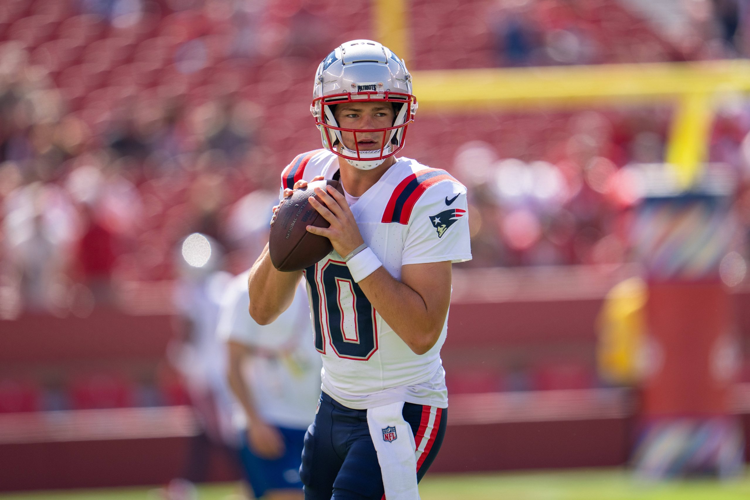 FootballR - NFL - Ein Footballspieler, Drake Maye, trägt ein weiß-rotes Trikot mit der Nummer 10 und einen Helm. Vor einer verschwommenen Stadionkulisse voller eifriger Zuschauer und hoch aufragender Torpfosten ist er bereit, den Ball zu werfen. Diese Beschreibung wurde automatisch generiert.