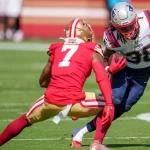 FootballR - NFL - Rhamondre Stevenson, in weiß-blauer Uniform, rennt mit dem Ball und wehrt einen Verteidiger in Rot und Gold mit der Nummer 7 ab. Sie stehen auf einem Rasenplatz und sind auf das Geschehen konzentriert. Der Hintergrund ist verschwommen und fängt die Intensität von Stevensons Kampf um einen Stammplatz ein. Diese Beschreibung wurde automatisch generiert.