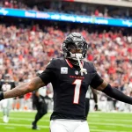 FootballR - NFL Free Agency 2025 - Ein Footballspieler in schwarzer Texans-Uniform mit der Nummer 1 steht mit ausgestreckten Armen da, nachdem er einen Touchdown erzielt hat. In seiner rechten Hand hält er einen Football, der an Stefon Diggs erinnert. Der unscharfe Stadionhintergrund ist mit jubelnden Fans und einem beleuchteten Bildschirm gefüllt. Diese Beschreibung wurde automatisch generiert.