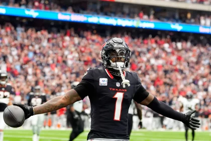 FootballR - NFL Free Agency 2025 - Ein Footballspieler in schwarzer Texans-Uniform mit der Nummer 1 steht mit ausgestreckten Armen da, nachdem er einen Touchdown erzielt hat. In seiner rechten Hand hält er einen Football, der an Stefon Diggs erinnert. Der unscharfe Stadionhintergrund ist mit jubelnden Fans und einem beleuchteten Bildschirm gefüllt. Diese Beschreibung wurde automatisch generiert.