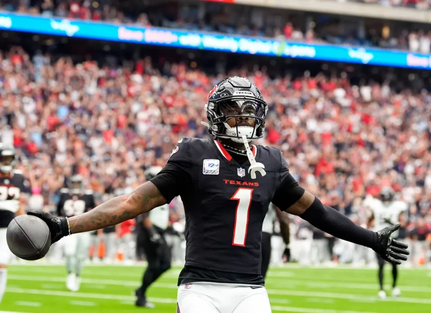 FootballR - NFL Free Agency 2025 - Ein Footballspieler in schwarzer Texans-Uniform mit der Nummer 1 steht mit ausgestreckten Armen da, nachdem er einen Touchdown erzielt hat. In seiner rechten Hand hält er einen Football, der an Stefon Diggs erinnert. Der unscharfe Stadionhintergrund ist mit jubelnden Fans und einem beleuchteten Bildschirm gefüllt. Diese Beschreibung wurde automatisch generiert.
