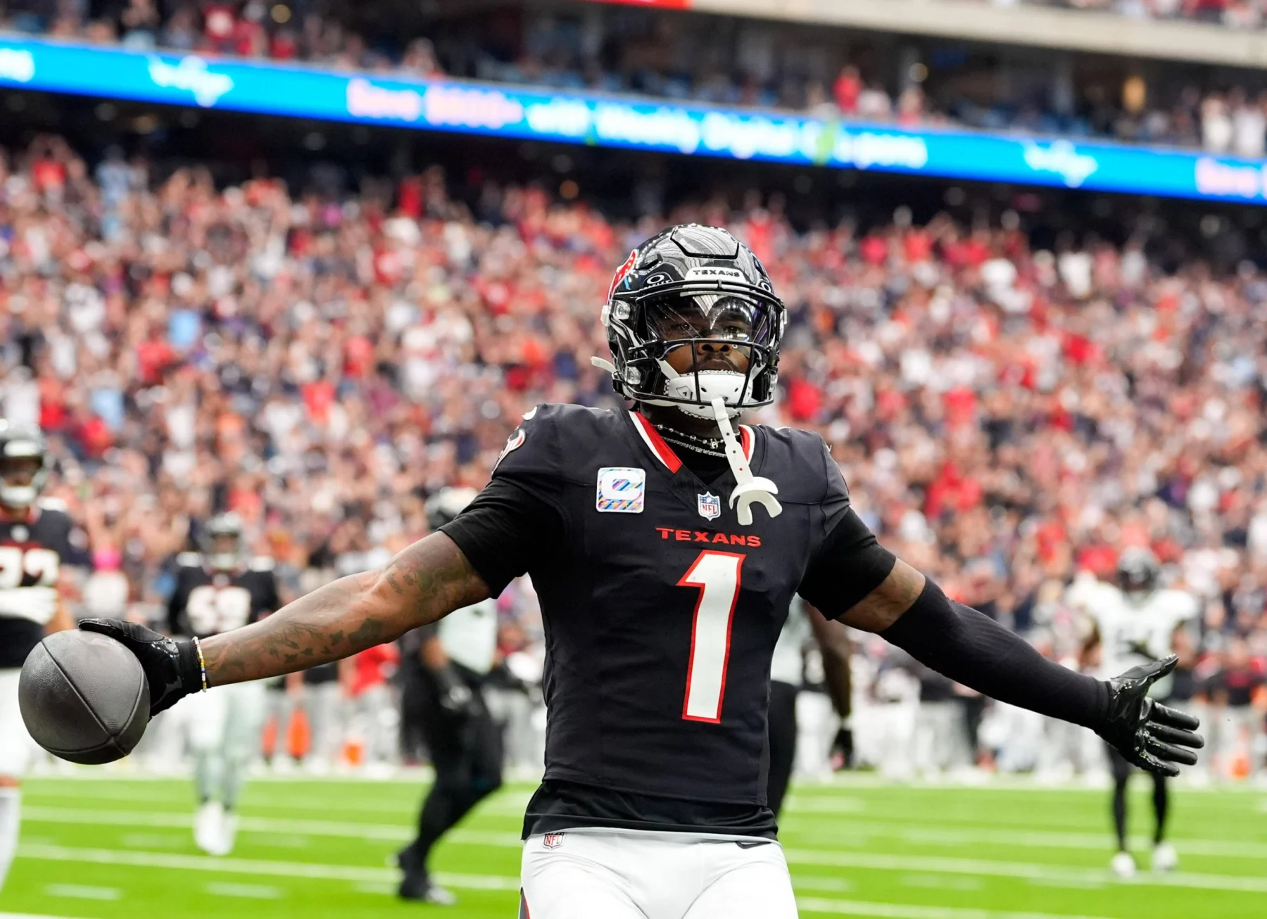 FootballR - NFL Free Agency 2025 - Ein Footballspieler in schwarzer Texans-Uniform mit der Nummer 1 steht mit ausgestreckten Armen da, nachdem er einen Touchdown erzielt hat. In seiner rechten Hand hält er einen Football, der an Stefon Diggs erinnert. Der unscharfe Stadionhintergrund ist mit jubelnden Fans und einem beleuchteten Bildschirm gefüllt. Diese Beschreibung wurde automatisch generiert.