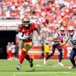 FootballR - NFL - Ein Footballspieler in rot-goldener Uniform mit Dreadlocks rennt mit dem Ball über ein Feld, verfolgt von Spielern in weiß-blauen Uniformen. Die Tribünen sind voller jubelnder Fans und bilden eine lebhafte Kulisse. Ein Schiedsrichter in schwarz-weißen Streifen beobachtet das Spiel von der Seitenlinie aus wie Fred Warner, der seinen nächsten Spielzug im Auge behält. Diese Beschreibung wurde automatisch generiert.