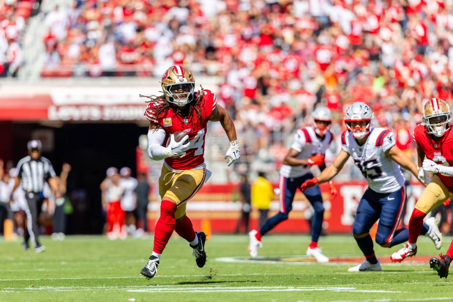 FootballR - NFL - Ein Footballspieler in rot-goldener Uniform mit Dreadlocks rennt mit dem Ball über ein Feld, verfolgt von Spielern in weiß-blauen Uniformen. Die Tribünen sind voller jubelnder Fans und bilden eine lebhafte Kulisse. Ein Schiedsrichter in schwarz-weißen Streifen beobachtet das Spiel von der Seitenlinie aus wie Fred Warner, der seinen nächsten Spielzug im Auge behält. Diese Beschreibung wurde automatisch generiert.