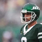 FootballR - DAZN NFL Programm - NFL - Ein Footballspieler in grün-weißer Jets-Uniform und Helm steht im Regen auf dem Spielfeld. Der Spieler hat einen ernsten Gesichtsausdruck und ist von der Hüfte aufwärts zu sehen, während Regentropfen um ihn herum fallen. Der Helm trägt das Jets-Logo, ein klares Visier über dem Gesicht und ist im DAZN NFL Programm zu sehen. Diese Beschreibung wurde automatisch generiert.