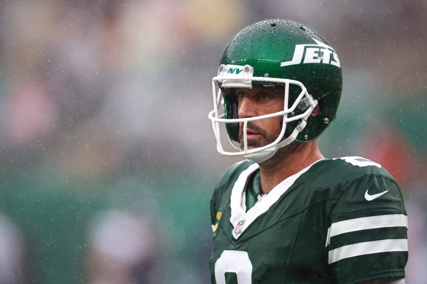 FootballR - DAZN NFL Programm - NFL - Ein Footballspieler in grün-weißer Jets-Uniform und Helm steht im Regen auf dem Spielfeld. Der Spieler hat einen ernsten Gesichtsausdruck und ist von der Hüfte aufwärts zu sehen, während Regentropfen um ihn herum fallen. Der Helm trägt das Jets-Logo, ein klares Visier über dem Gesicht und ist im DAZN NFL Programm zu sehen. Diese Beschreibung wurde automatisch generiert.