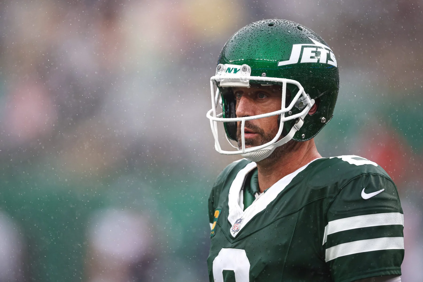 FootballR - DAZN NFL Programm - NFL - Ein Footballspieler in grün-weißer Jets-Uniform und Helm steht im Regen auf dem Spielfeld. Der Spieler hat einen ernsten Gesichtsausdruck und ist von der Hüfte aufwärts zu sehen, während Regentropfen um ihn herum fallen. Der Helm trägt das Jets-Logo, ein klares Visier über dem Gesicht und ist im DAZN NFL Programm zu sehen. Diese Beschreibung wurde automatisch generiert.
