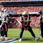 FootballR - NFL - Ein Footballspieler in einer rot-weißen Patriots-Uniform macht auf dem Spielfeld eine Siegespose. Teamkollegen in passenden Uniformen, darunter Jabrill Peppers, stehen in der Nähe. Ein Offizieller in einem schwarz-weiß gestreiften Hemd schaut zu. Das Stadion ist voller Fans und helles Sonnenlicht erhellt die Szene. Diese Beschreibung wurde automatisch generiert.