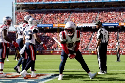FootballR - NFL - Ein Footballspieler in einer rot-weißen Patriots-Uniform macht auf dem Spielfeld eine Siegespose. Teamkollegen in passenden Uniformen, darunter Jabrill Peppers, stehen in der Nähe. Ein Offizieller in einem schwarz-weiß gestreiften Hemd schaut zu. Das Stadion ist voller Fans und helles Sonnenlicht erhellt die Szene. Diese Beschreibung wurde automatisch generiert.