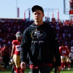 FootballR - NFL - Ein Mann mit schwarzem Kapuzenpullover und schwarzer Baseballkappe läuft über ein Footballfeld. Auf dem Kapuzenpullover ist das Logo „Crucial Catch“ zu sehen. Hinter ihm sind Footballspieler in rot-goldenen Uniformen zu sehen, darunter Christian McCaffrey, und die Tribünen des Stadions sind voller Zuschauer. Der Himmel ist klar und hell. Diese Beschreibung wurde automatisch generiert.