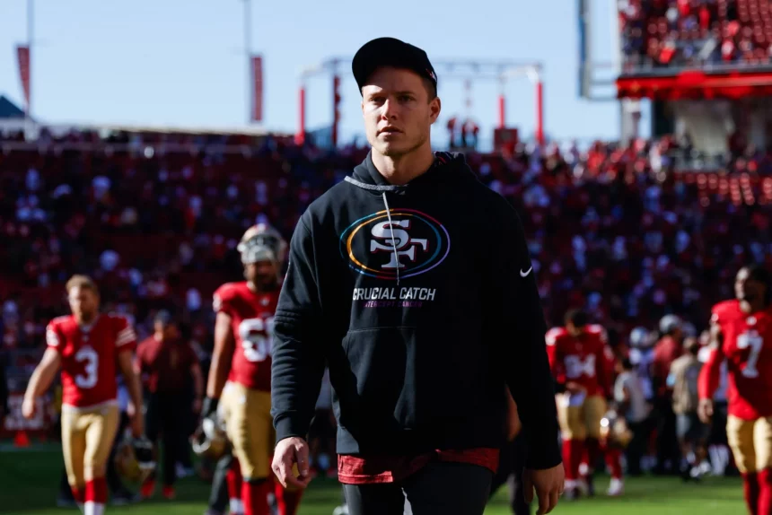 FootballR - NFL - Ein Mann mit schwarzem Kapuzenpullover und schwarzer Baseballkappe läuft über ein Footballfeld. Auf dem Kapuzenpullover ist das Logo „Crucial Catch“ zu sehen. Hinter ihm sind Footballspieler in rot-goldenen Uniformen zu sehen, darunter Christian McCaffrey, und die Tribünen des Stadions sind voller Zuschauer. Der Himmel ist klar und hell. Diese Beschreibung wurde automatisch generiert.