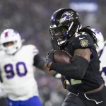 FootballR - NFL heute im TV - Ein Footballspieler der Baltimore Ravens, der ein schwarzes Trikot mit der Nummer 22 trägt, läuft mit dem Ball während eines Spiels gegen die Buffalo Bills. Verteidiger in weißen Trikots verfolgen ihn auf einem regennassen Feld unter Stadionbeleuchtung. Dieses spannende Spiel könnte das NFL Power Ranking in dieser Saison beeinflussen. Diese Beschreibung wurde automatisch generiert.