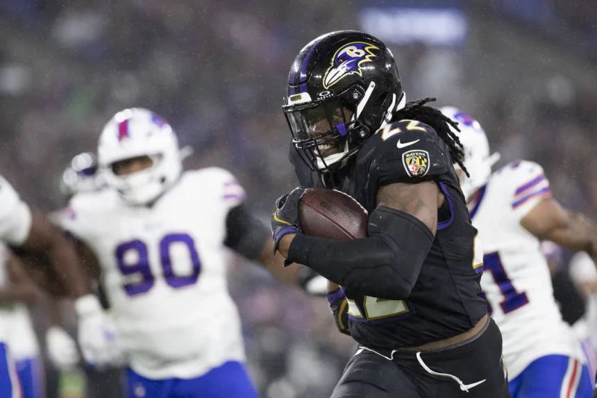 FootballR - NFL heute im TV - Ein Footballspieler der Baltimore Ravens, der ein schwarzes Trikot mit der Nummer 22 trägt, läuft mit dem Ball während eines Spiels gegen die Buffalo Bills. Verteidiger in weißen Trikots verfolgen ihn auf einem regennassen Feld unter Stadionbeleuchtung. Dieses spannende Spiel könnte das NFL Power Ranking in dieser Saison beeinflussen. Diese Beschreibung wurde automatisch generiert.