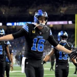 Week 7 Predictions - NFL - Jared Goff, der ein Trikot mit der Nummer 16 der Detroit Lions trägt, streckt seine Arme aus, um einem Teamkollegen ein High-Five zu geben. Er steht auf einem Footballfeld, andere Spieler und ein unscharfer Stadionhintergrund sind zu sehen. Der Spieler trägt schwarze Kleidung mit blauen Akzenten und einen Helm. Diese Beschreibung wurde automatisch generiert.
