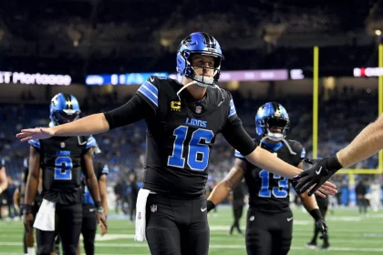 Week 7 Predictions - NFL - Jared Goff, der ein Trikot mit der Nummer 16 der Detroit Lions trägt, streckt seine Arme aus, um einem Teamkollegen ein High-Five zu geben. Er steht auf einem Footballfeld, andere Spieler und ein unscharfer Stadionhintergrund sind zu sehen. Der Spieler trägt schwarze Kleidung mit blauen Akzenten und einen Helm. Diese Beschreibung wurde automatisch generiert.