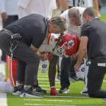 FootballR - NFL - Ein Footballspieler in einem weißen Chiefs-Trikot und einem roten Helm - Rashee Rice - kniet auf dem Feld, gestützt von zwei Mitarbeitern in schwarzen Hemden und grauen Hosen. Sie scheinen seinen Zustand zu überprüfen, wobei einer der Mitarbeiter ein Funkgerät hält. Im Hintergrund sind weitere Teammitglieder in ähnlichen Uniformen zu sehen. Diese Beschreibung wurde automatisch generiert.