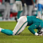 FootballR - NFL - Jaelan Phillips - Ein Footballspieler in einer blaugrünen und weißen Dolphins-Uniform kniet mit gesenktem Kopf auf dem Feld und wirkt erschöpft oder frustriert. Er trägt einen weißen Helm mit einem orangefarbenen Logo. Der Hintergrund ist verschwommen und zeigt andere Spieler und die Seitenlinie. Der Rasen ist grün und vom Spiel abgenutzt. Diese Beschreibung wurde automatisch generiert.