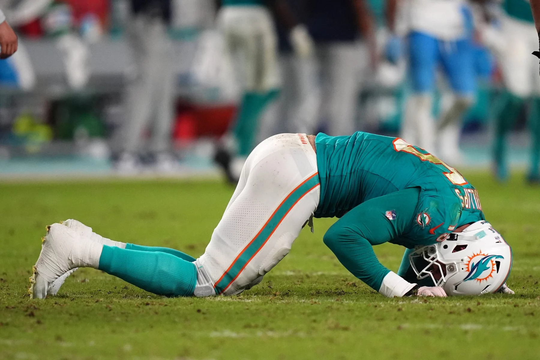 FootballR - NFL - Jaelan Phillips - Ein Footballspieler in einer blaugrünen und weißen Dolphins-Uniform kniet mit gesenktem Kopf auf dem Feld und wirkt erschöpft oder frustriert. Er trägt einen weißen Helm mit einem orangefarbenen Logo. Der Hintergrund ist verschwommen und zeigt andere Spieler und die Seitenlinie. Der Rasen ist grün und vom Spiel abgenutzt. Diese Beschreibung wurde automatisch generiert.