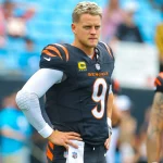 FootballR - NFL - Ein Footballspieler in einer schwarz-orangen Bengals-Uniform steht auf dem Feld und trägt stolz das Trikot mit der Nummer 9. Mit hellem Haar und einem gepolsterten weißen Ärmel ähnelt er Joe Burrow. Ein anderer Spieler ist teilweise im Hintergrund zu sehen, während verschwommene Zuschauer dieses Schlüsselspiel gegen die Ravens verfolgen. Diese Beschreibung wurde automatisch generiert.