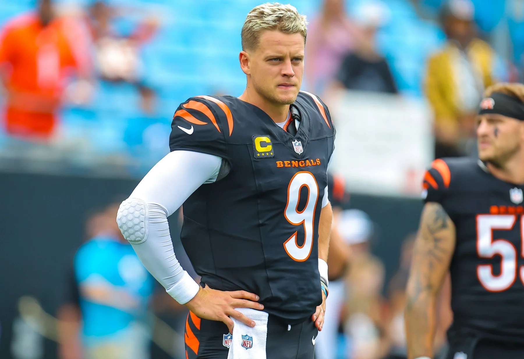 FootballR - NFL - Ein Footballspieler in einer schwarz-orangen Bengals-Uniform steht auf dem Feld und trägt stolz das Trikot mit der Nummer 9. Mit hellem Haar und einem gepolsterten weißen Ärmel ähnelt er Joe Burrow. Ein anderer Spieler ist teilweise im Hintergrund zu sehen, während verschwommene Zuschauer dieses Schlüsselspiel gegen die Ravens verfolgen. Diese Beschreibung wurde automatisch generiert.