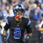 Ein Footballspieler, Jameson Williams, trägt ein schwarzes Trikot mit blauen und silbernen Akzenten, Nummer 9, von den Detroit Lions. Er hat einen Helm mit Visier und einen roten Mundschutz. Im Hintergrund ist ein verschwommenes Stadion voller Zuschauer und ein Offizieller in schwarz-weißen Streifen zu sehen. Diese Beschreibung wurde mit der FootballR KI automatisch generiert.