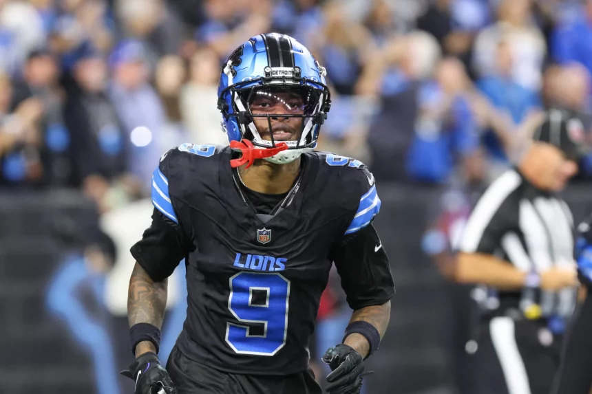 Ein Footballspieler, Jameson Williams, trägt ein schwarzes Trikot mit blauen und silbernen Akzenten, Nummer 9, von den Detroit Lions. Er hat einen Helm mit Visier und einen roten Mundschutz. Im Hintergrund ist ein verschwommenes Stadion voller Zuschauer und ein Offizieller in schwarz-weißen Streifen zu sehen. Diese Beschreibung wurde mit der FootballR KI automatisch generiert.