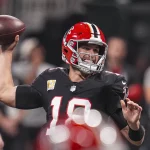 FootballR - NFL Fantasy Football Week 11 - Ein Footballspieler in schwarzer Uniform mit roten und weißen Akzenten und dem Emblem der Falcons - Kirk Cousins - bereitet sich auf den Einwurf vor, der ein spannendes Spiel in der Verlängerung verspricht. Im Hintergrund verschwimmen Zuschauer und Spieler. Seine konzentrierte Intensität ist unter seinem Kapitänsabzeichen spürbar. Diese Beschreibung wurde automatisch generiert.