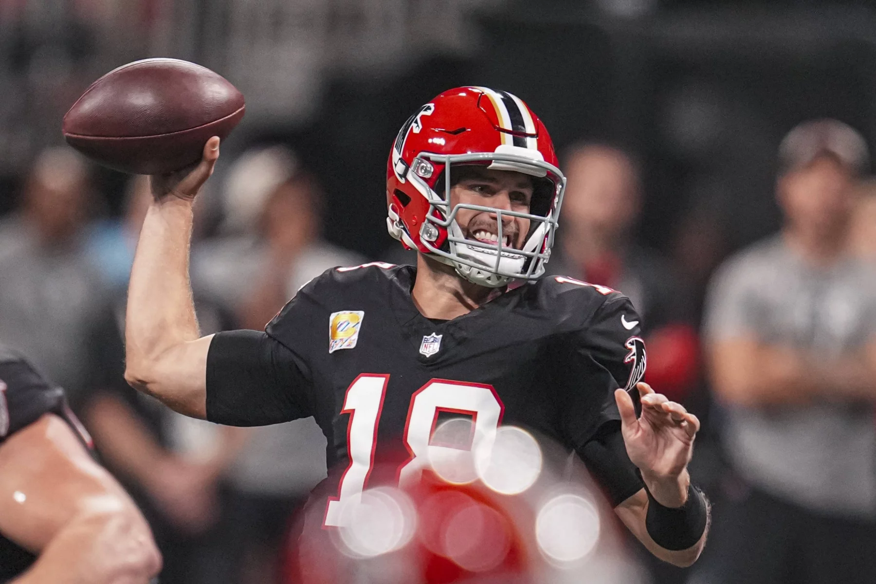 FootballR - NFL Fantasy Football Week 11 - Ein Footballspieler in schwarzer Uniform mit roten und weißen Akzenten und dem Emblem der Falcons - Kirk Cousins - bereitet sich auf den Einwurf vor, der ein spannendes Spiel in der Verlängerung verspricht. Im Hintergrund verschwimmen Zuschauer und Spieler. Seine konzentrierte Intensität ist unter seinem Kapitänsabzeichen spürbar. Diese Beschreibung wurde automatisch generiert.