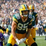 FootballR - NFL - Tucker Kraft in grün-gelber Uniform mit der Nummer 85 feiert in Week 6 auf dem Feld. Er trägt einen Helm, ballt die Fäuste und schreit. Im Hintergrund sind andere Spieler und Offizielle zu sehen, während Fans die Sitze im Stadion unter einem klaren Himmel füllen. Diese Beschreibung wurde automatisch generiert.