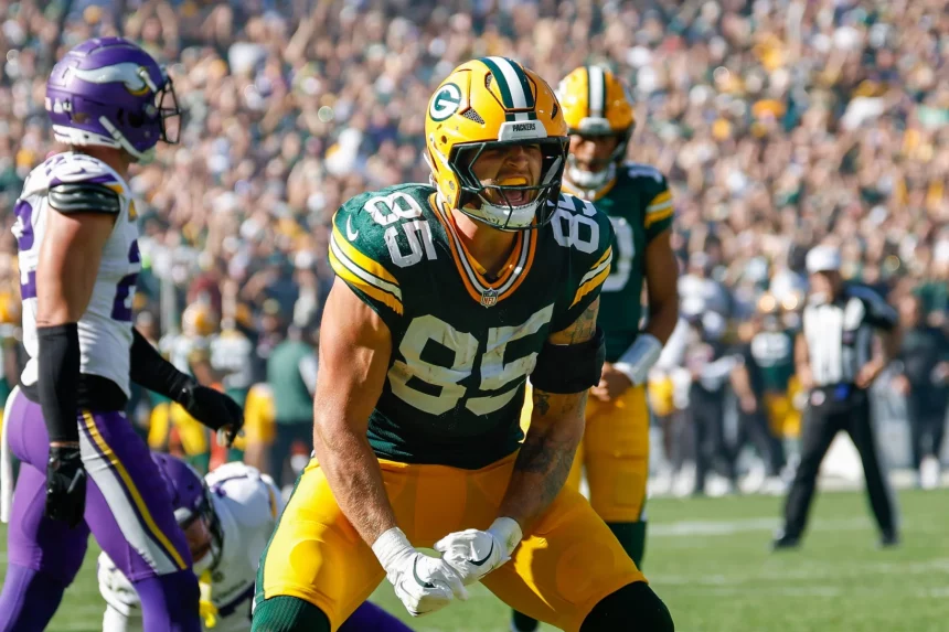 FootballR - NFL - Tucker Kraft in grün-gelber Uniform mit der Nummer 85 feiert in Week 6 auf dem Feld. Er trägt einen Helm, ballt die Fäuste und schreit. Im Hintergrund sind andere Spieler und Offizielle zu sehen, während Fans die Sitze im Stadion unter einem klaren Himmel füllen. Diese Beschreibung wurde automatisch generiert.