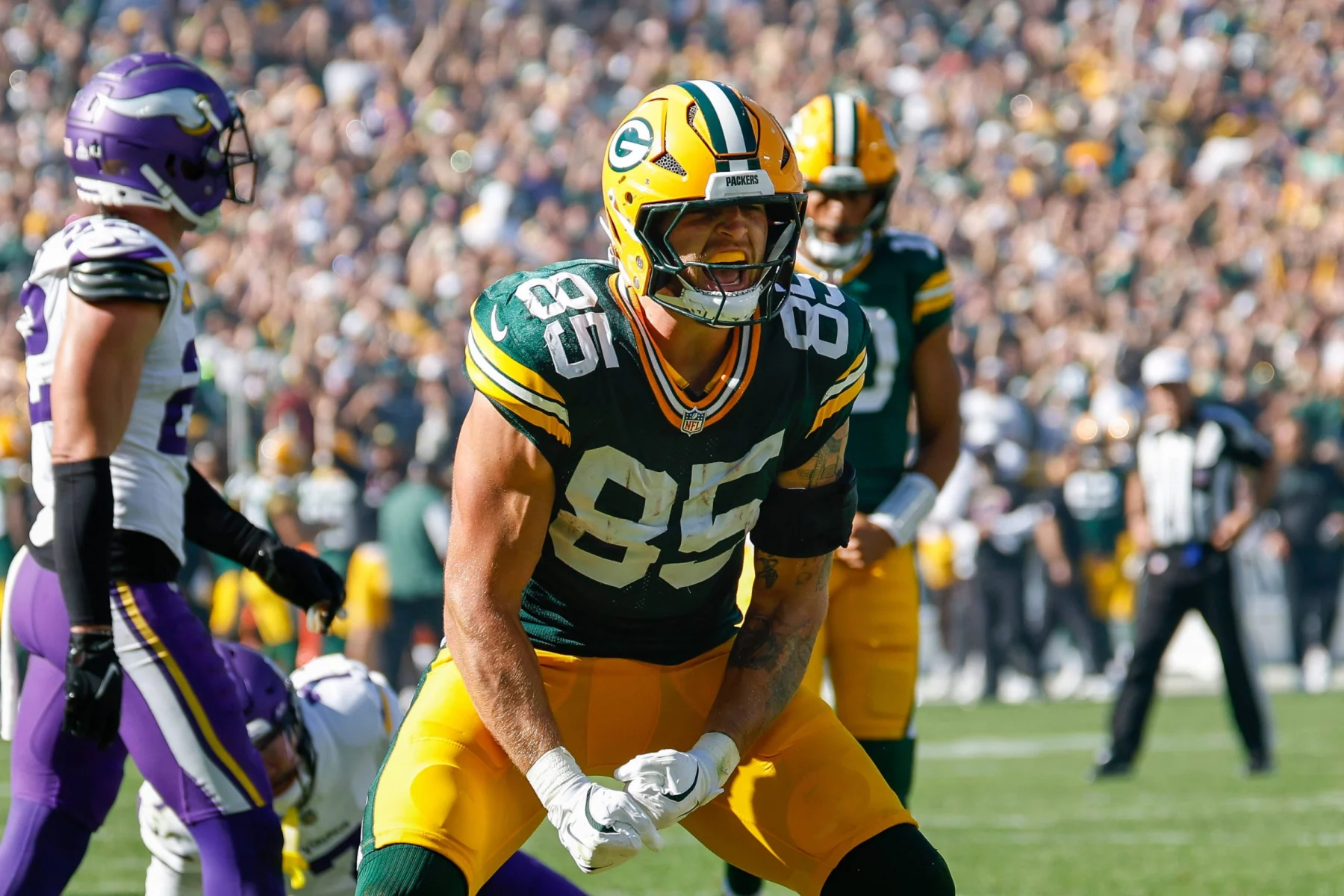 FootballR - NFL - Tucker Kraft in grün-gelber Uniform mit der Nummer 85 feiert in Week 6 auf dem Feld. Er trägt einen Helm, ballt die Fäuste und schreit. Im Hintergrund sind andere Spieler und Offizielle zu sehen, während Fans die Sitze im Stadion unter einem klaren Himmel füllen. Diese Beschreibung wurde automatisch generiert.